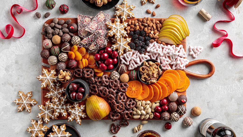 Photo of a holiday dessert board with an assortment of chocolate covered pretzels, cookies, dried fruit slices, sliced pears and mangos, nuts and chocolates
