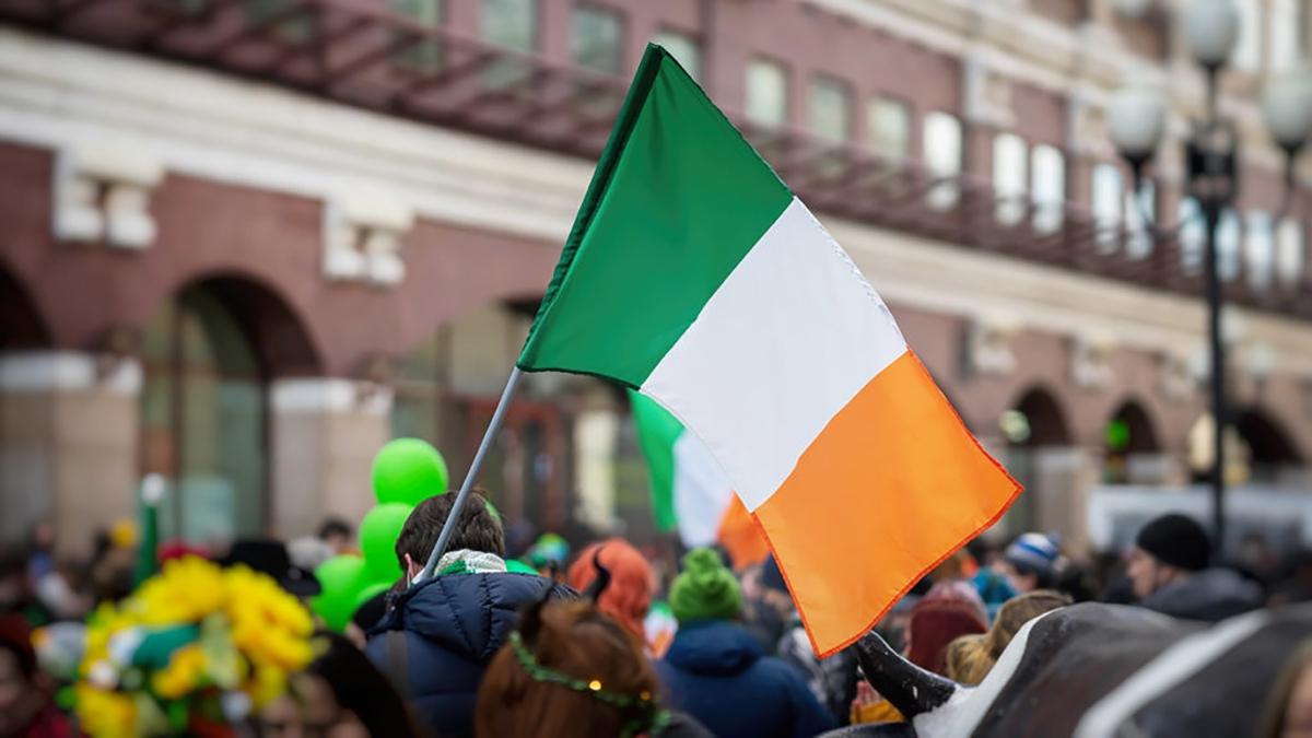 National Flag of Ireland close up in hands of man on background of crowd people in city street during celebration of St. Patrick's Day