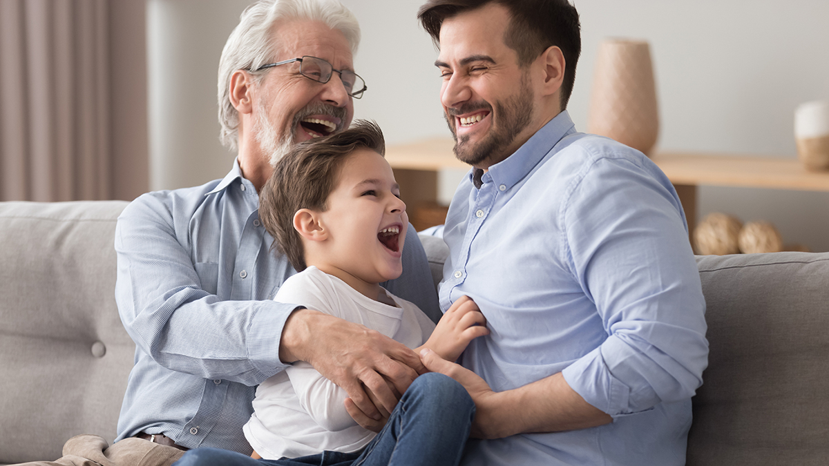 A photo of celebrate father's day with a grandfather, son and grandson sitting on a couch and laughing.