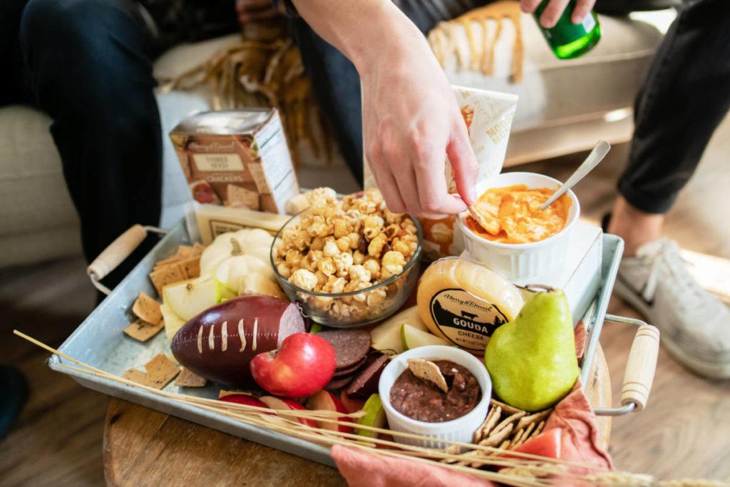 tailgate party image   snacks spread out on a board with a hand dipping a pretzel into a bowl of dip