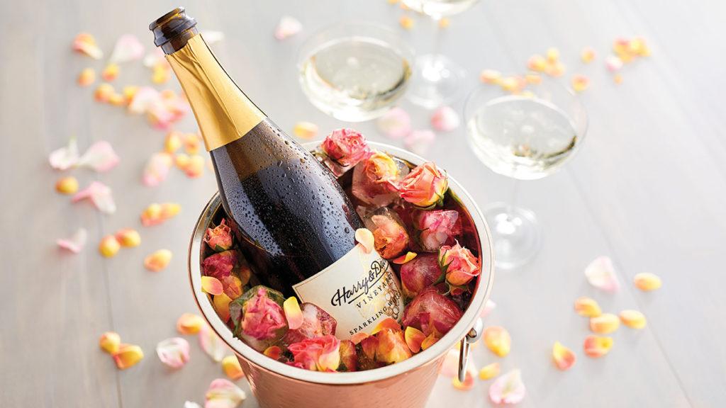 A photo of sparkling wine with a bottle of wine in a bucket full of flowers with two glasses of wine in the background.