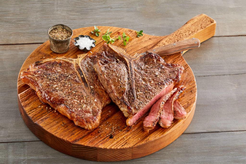 A photo of cuts of beef with a cutting board supporting two cooked t bone steaks.