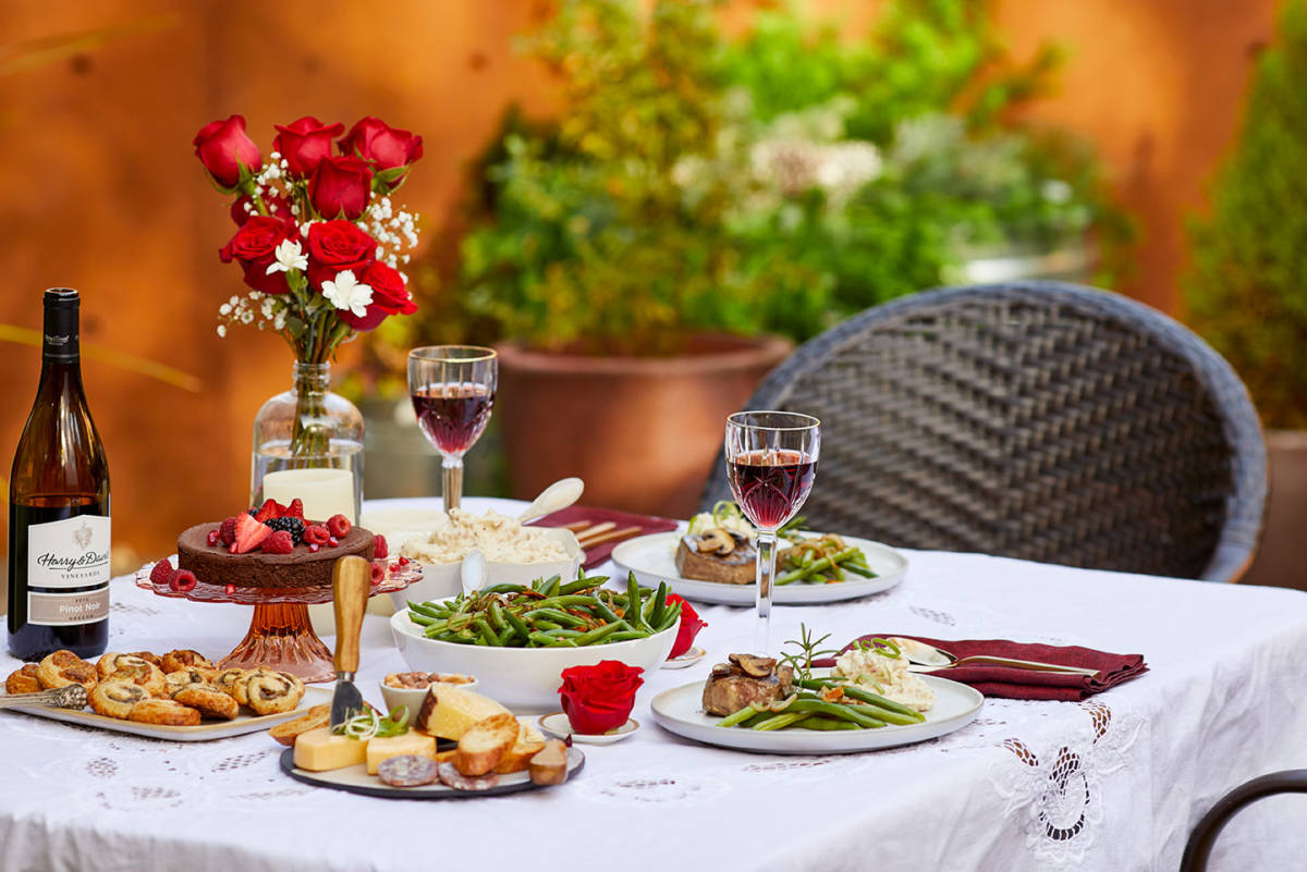 A photo of valentine's day date ideas with a table outside supporting a gourmet meal with wine and flowers.