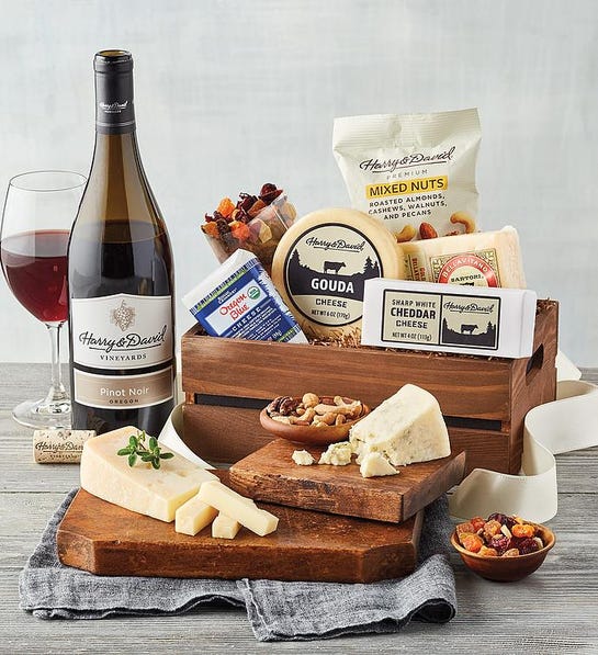 A photo of birthday gift ideas with a box full of cheese and snacks next to a bottle of wine and full glass with a cutting board holding more cheese and nuts in front