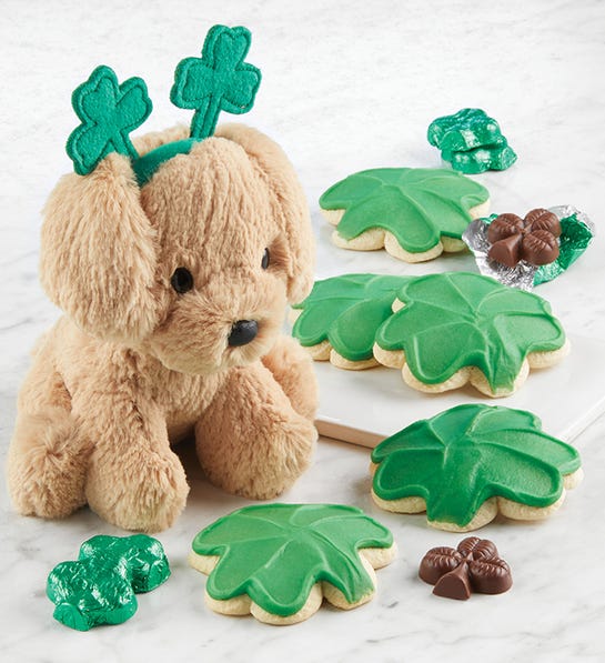 A photo of St Patricks Day with a stuffed dog wearing a headband that has two shamrocks on it surrounded by shamrock shaped cookies chocolate