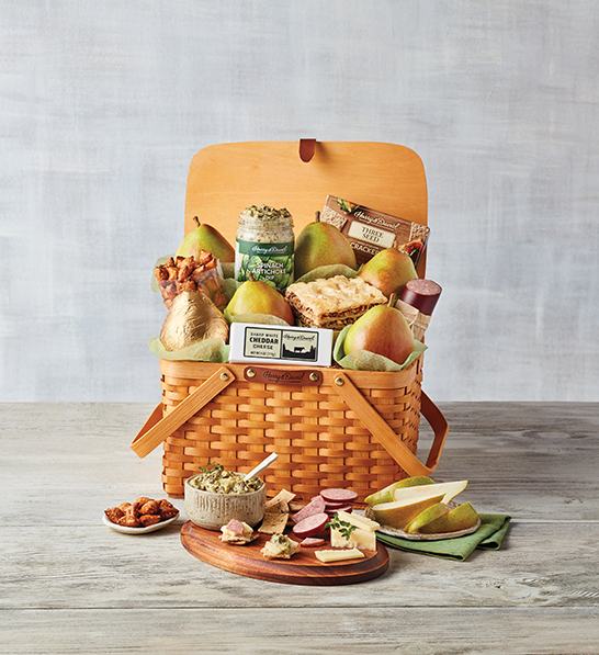 A photo of gifts for women with a picnic basket full of fruit crackers cheese meat and spreads with the same ingredients on a plate in front of the basket