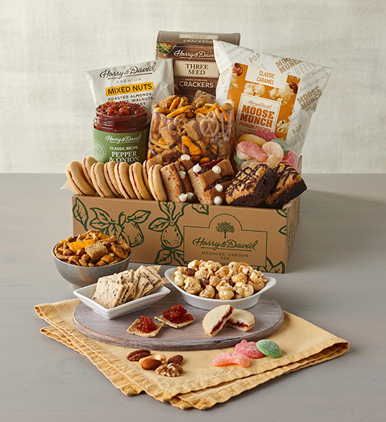 A photo of gifts for women with a box full of sweet and salty snacks with the same items on several plates and bowls in front of the box