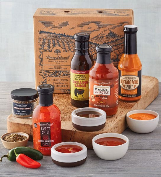 A photo of gifts for men with four jars of grilling sauces on a wooden cutting board with four ramekins full of the same sauces next to the them and two other ramekins full of spices