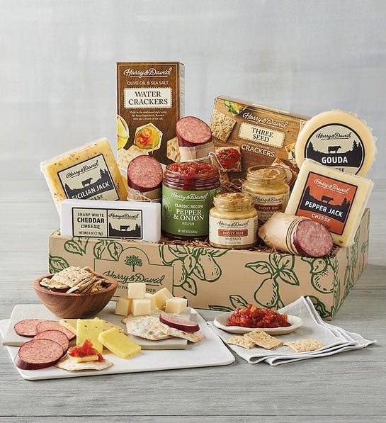 A photo of gifts for men with a box full of cured meats cheeses and spreads with the same ingredients on a cutting board and plates in front of the box