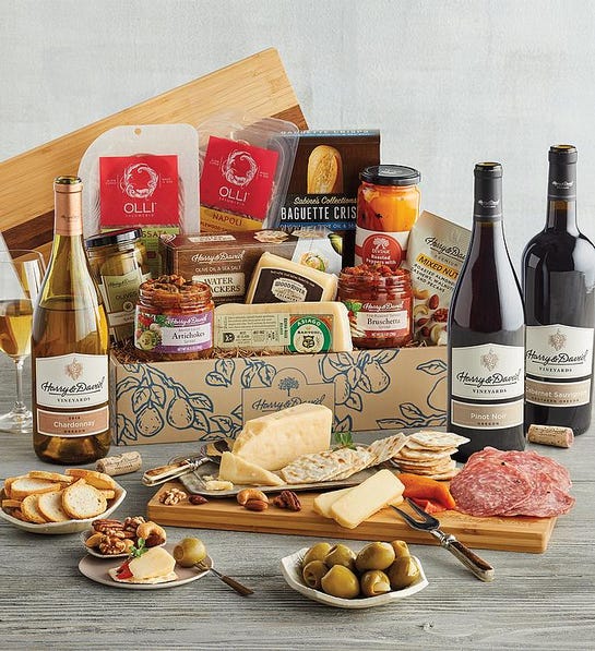 A photo of gifts for men with a box overflowing with meats cheeses spreads and olives surrounded by a cutting board full of the same ingredients and four bottles of wine