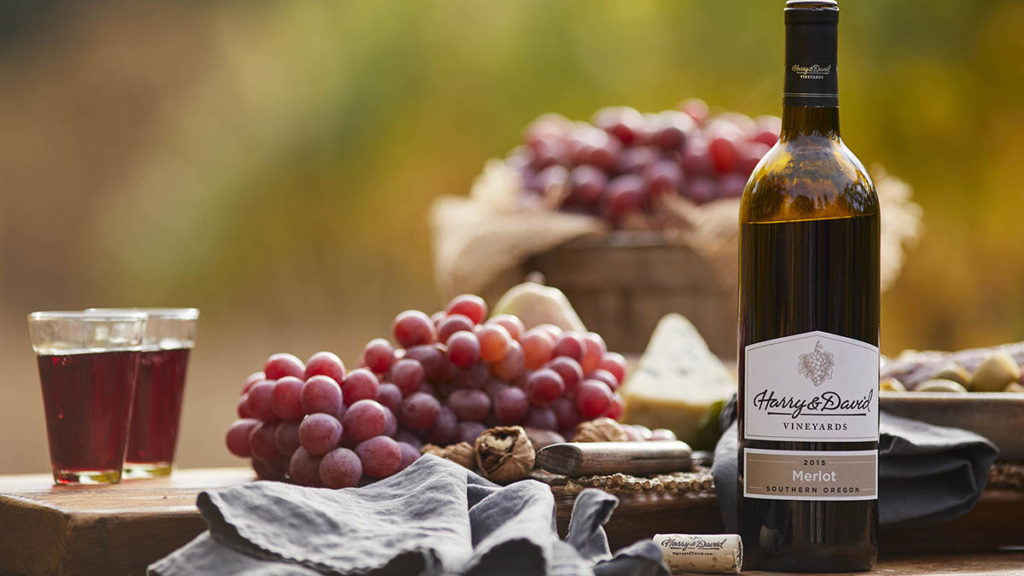 Wine pronunciation with a bottle of red wine on a table next to a bunch of grapes and two glasses full of wine.