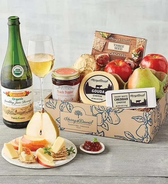 A photo of gifts for new moms with a crate of cheese fruit and crackers next to a bottle of sparkling juice and a plate full of apples and cheese