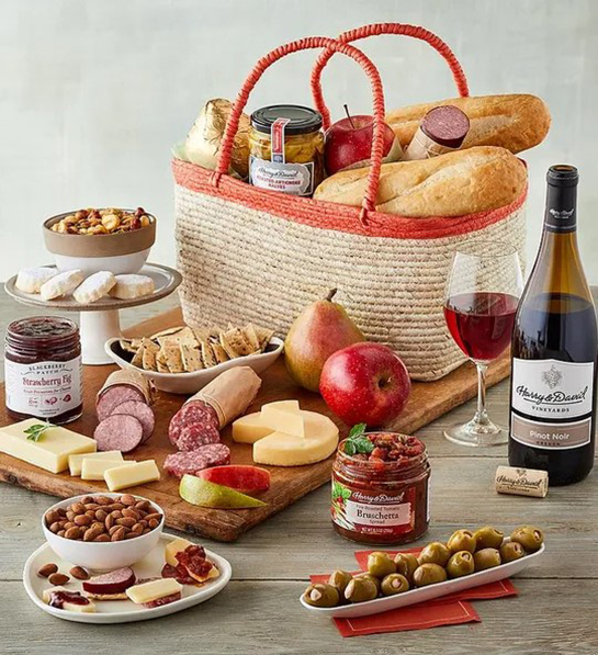 A photo of picnic with a basket full of cheese bread and other snacks surrounded by the same ingredients and a bottle of wine