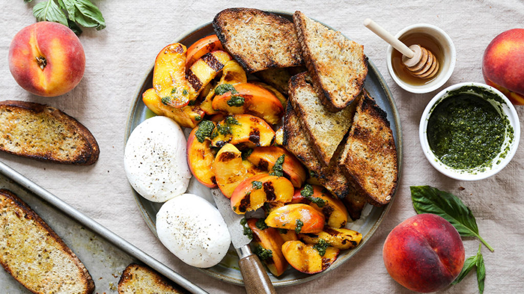 Photo of how to grill with a plate full of grilled peaches toasted bread and burrata with a bowl of pesto and a bowl of honey next to the plate