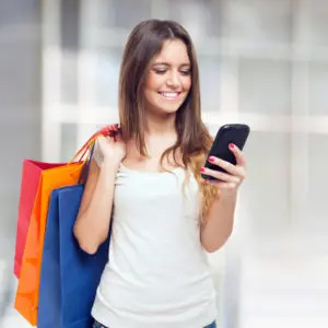 Office superstar smiling at her phone with several shopping bags slung over her shoulder.